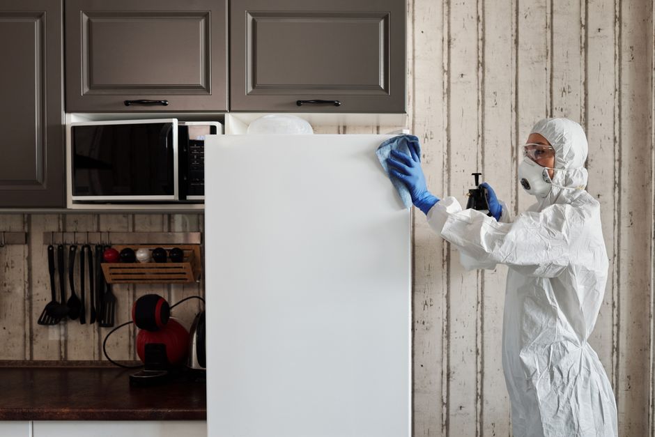 Close-up of a person dressed in white protective clothing and blue gloves, engaged in surface sanitisation inside a home or office environment. The individual holds a black spray gun connected to a wh